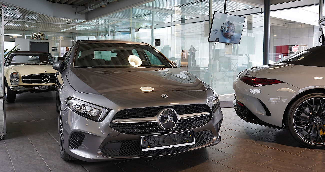 Firmen Schild Innenaufnahme des Autohauses zeigt im Vordergrund eine graue A-Klasse, dahinter bzw. daneben einen cremefarbenen SL-Oldtimer und einen weißen SL 63 AMG